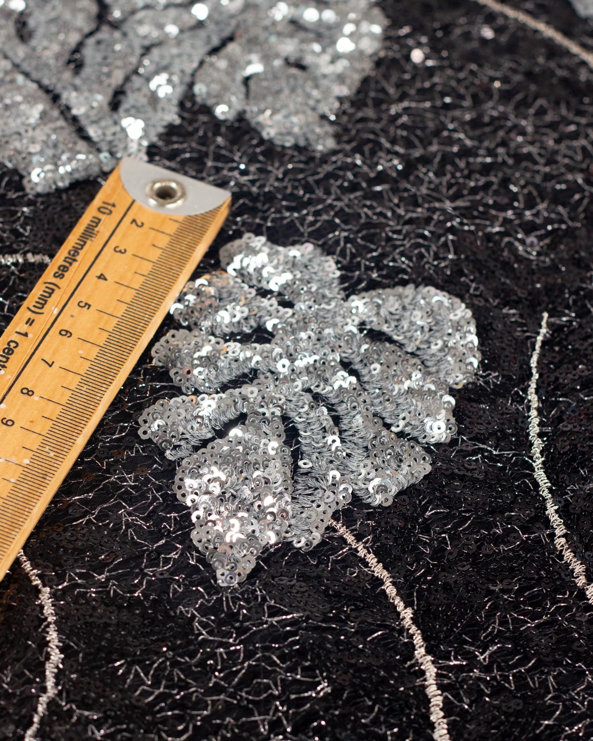 A close up of a silver monstera leaf applique made of densely packed silver sequins on a black cobweb tulle fabric with silver embroidery forming the stems and with a wooden ruler for scale.