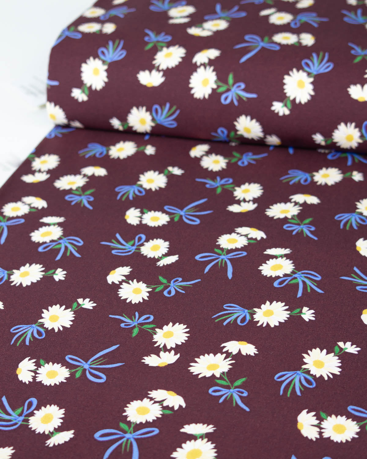 A roll of cotton canvas fabric with a dark burgundy background patterned with white daisies, blue bows, and green stems, shown partially unrolled.