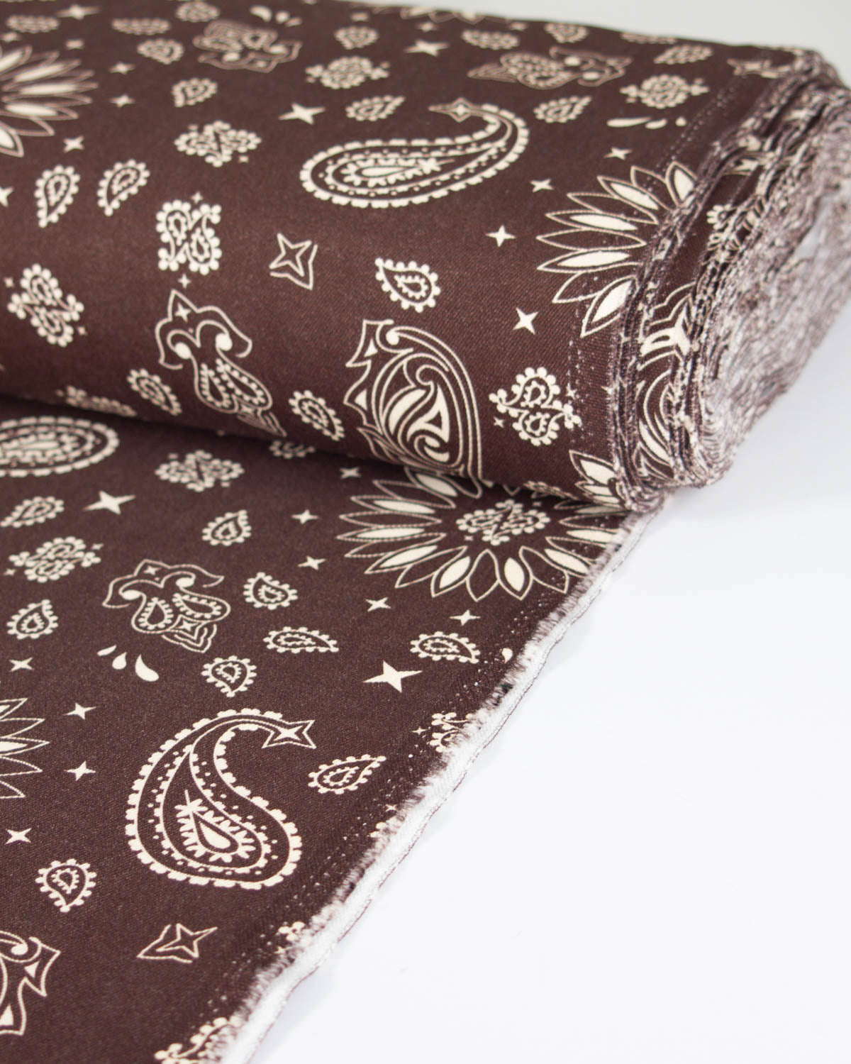 A roll of dark brown cotton denim fabric printed with a cream paisley and floral bandana pattern, shown on a white background.