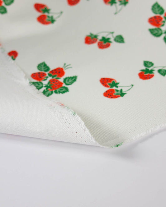 White cotton denim fabric with a repeating strawberry print in red and green, folded over to show the woven texture and raw edge.
