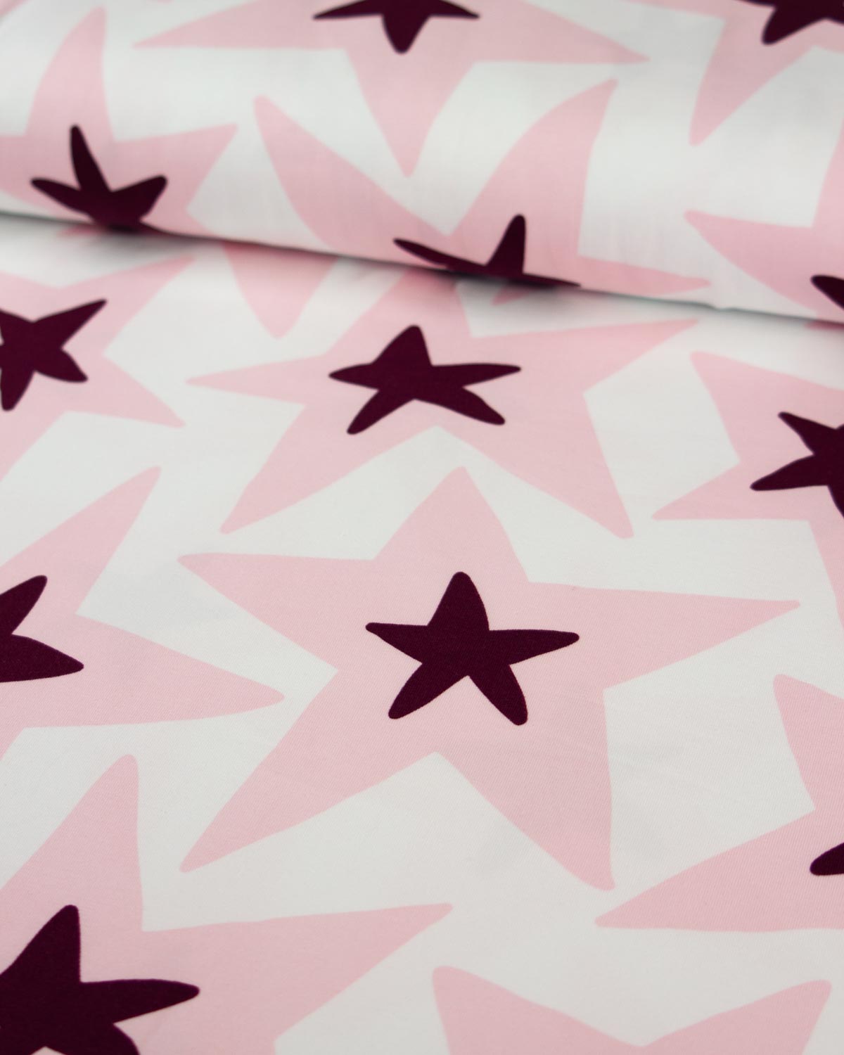 A close-up of patterned cotton jersey fabric with large light pink stars on a white background and darker plum-purple star centers.