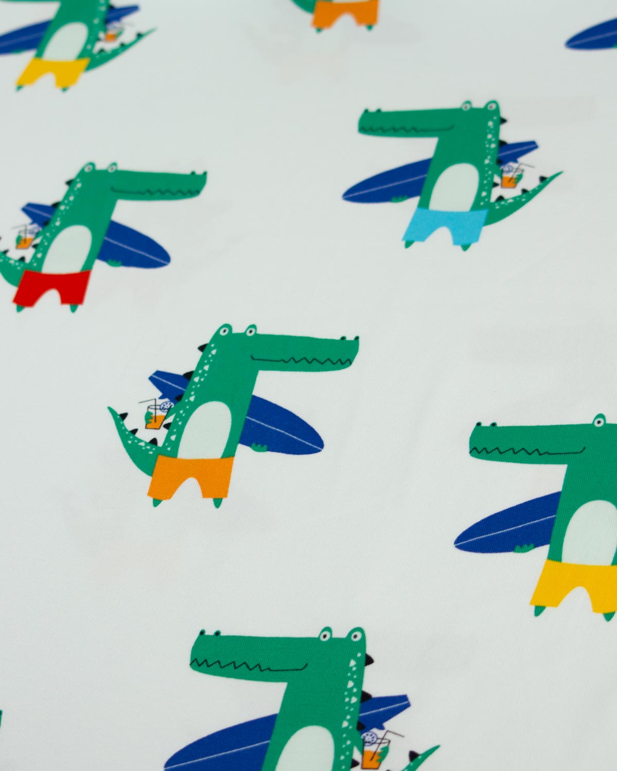A close-up of a white cotton jersey fabric printed with a repeating pattern of green cartoon crocodiles flying on blue surfboards, with small multicolor accents.