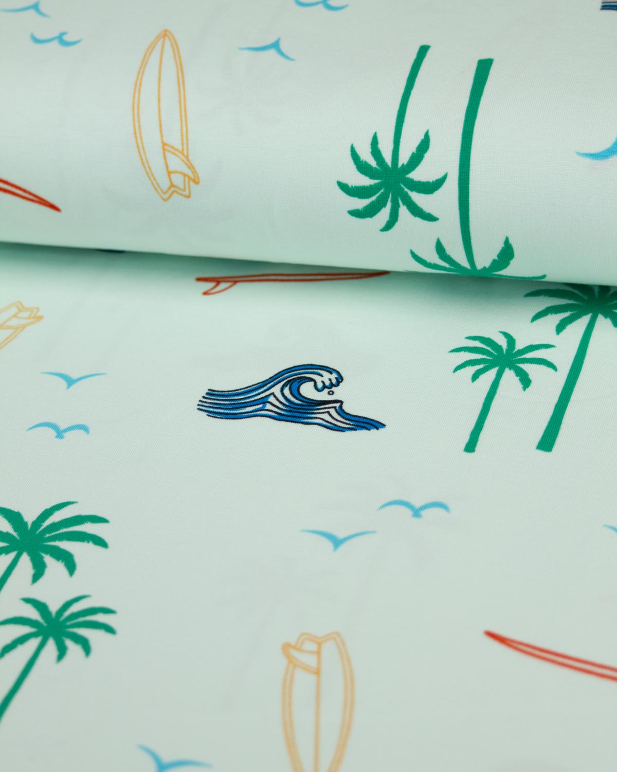 A close-up of a light mint green cotton jersey fabric with a tropical beach print featuring green palm trees, blue waves, and yellow and orange surfboards.