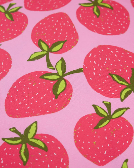 A close-up of a pink cotton jersey fabric pattern featuring illustrated red strawberries with green leaves and small white seed details.
