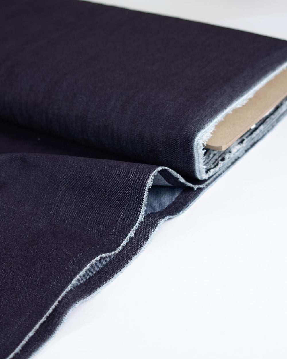 A close-up of dark indigo denim fabric, folded and draped to show its texture and raw, frayed edges against a clean white background.