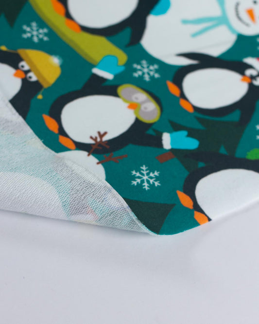 A close-up of a folded fabric featuring a penguin pattern on a teal background. The fabric is shown layered over a white surface, displaying its sheer, semi-transparent texture.