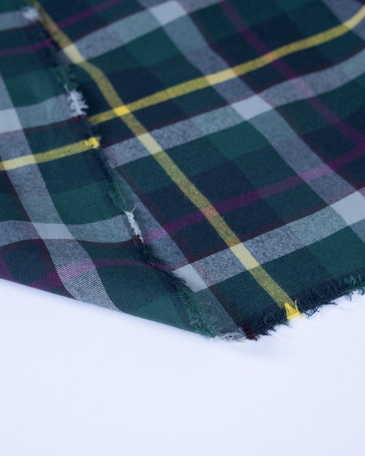 A close-up of a folded plaid fabric with a tartan pattern in dark green, navy, purple, gray, and yellow, showing a frayed edge against a plain white background.