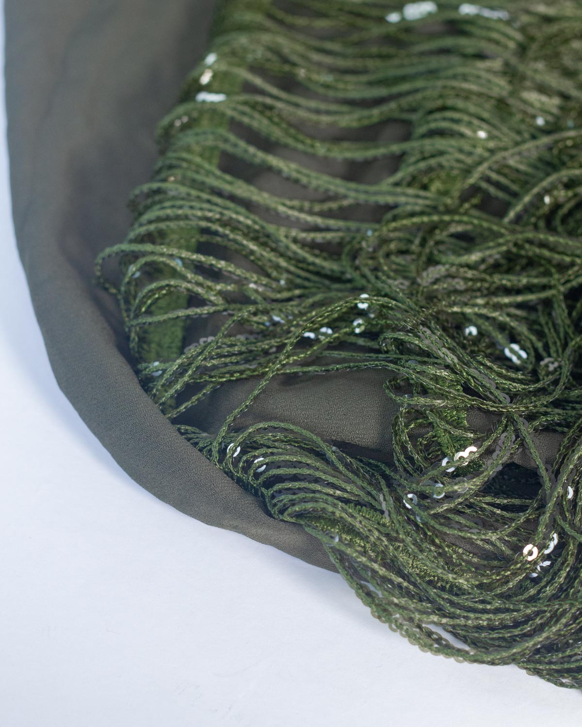 Close-up of an olive green polyester fabric with layered fringe sequin strands and scattered small silver sequins, laid on a white background.