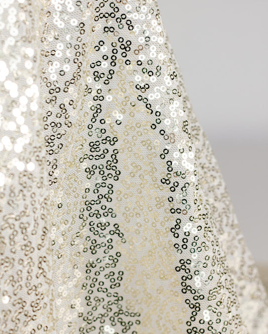 A close-up of draped sequined tulle fabric with shimmering champagne gold sequins on a white base, against a neutral gray background.