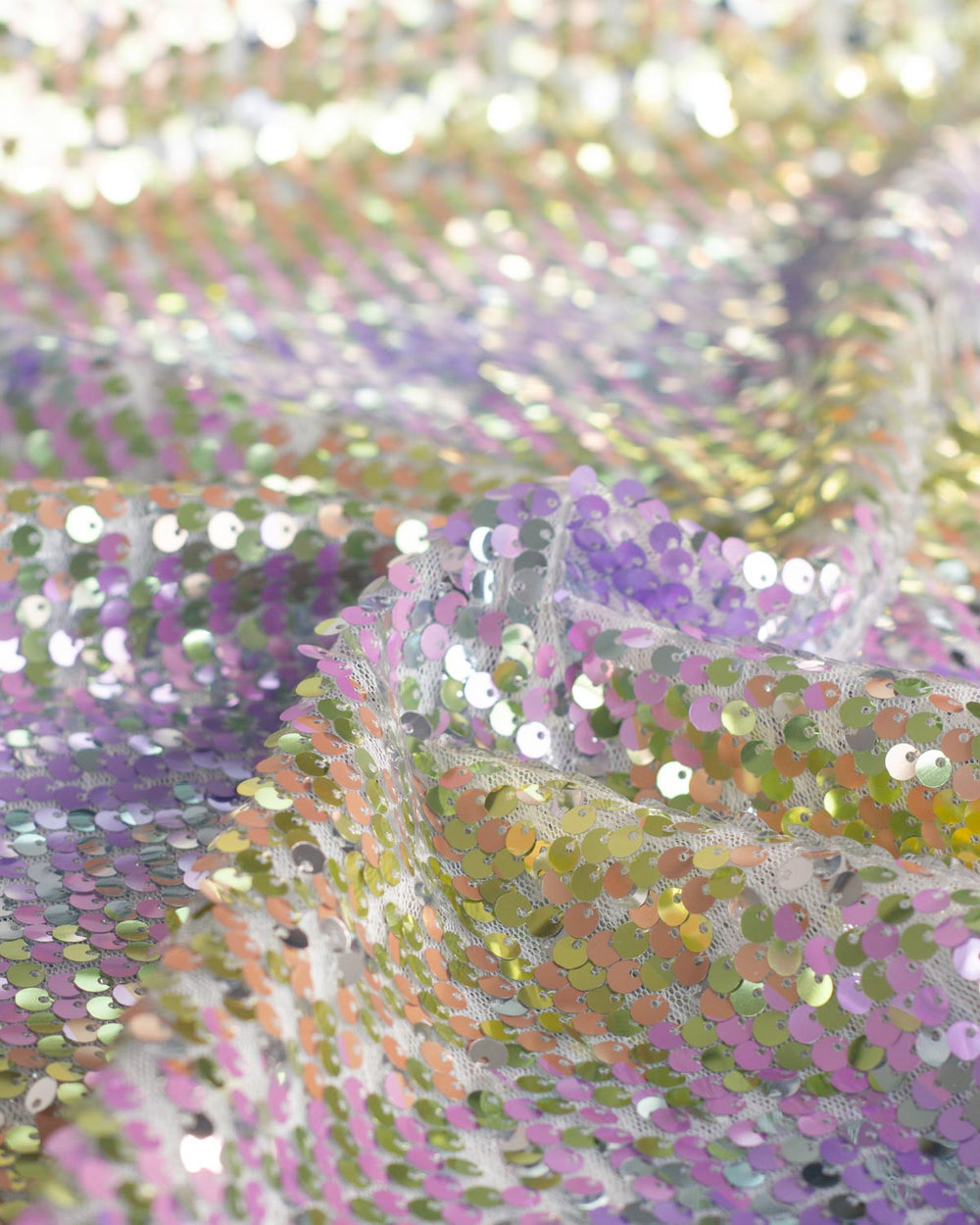 Close-up of iridescent sequin tulle fabric with shimmering pastel tones including lavender, lilac, peach, and gold, draped in soft folds.