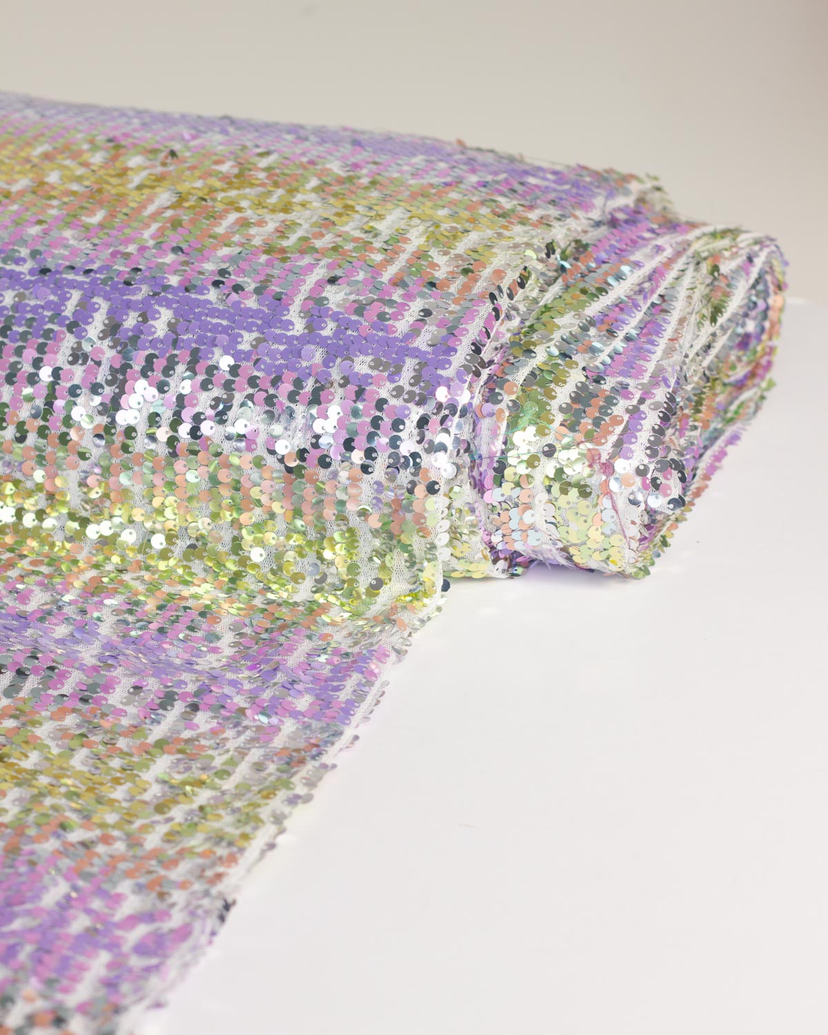 A close-up of a roll of iridescent sequin fabric showing a pastel prism gradient of purple, green, and gold on a plain white background.