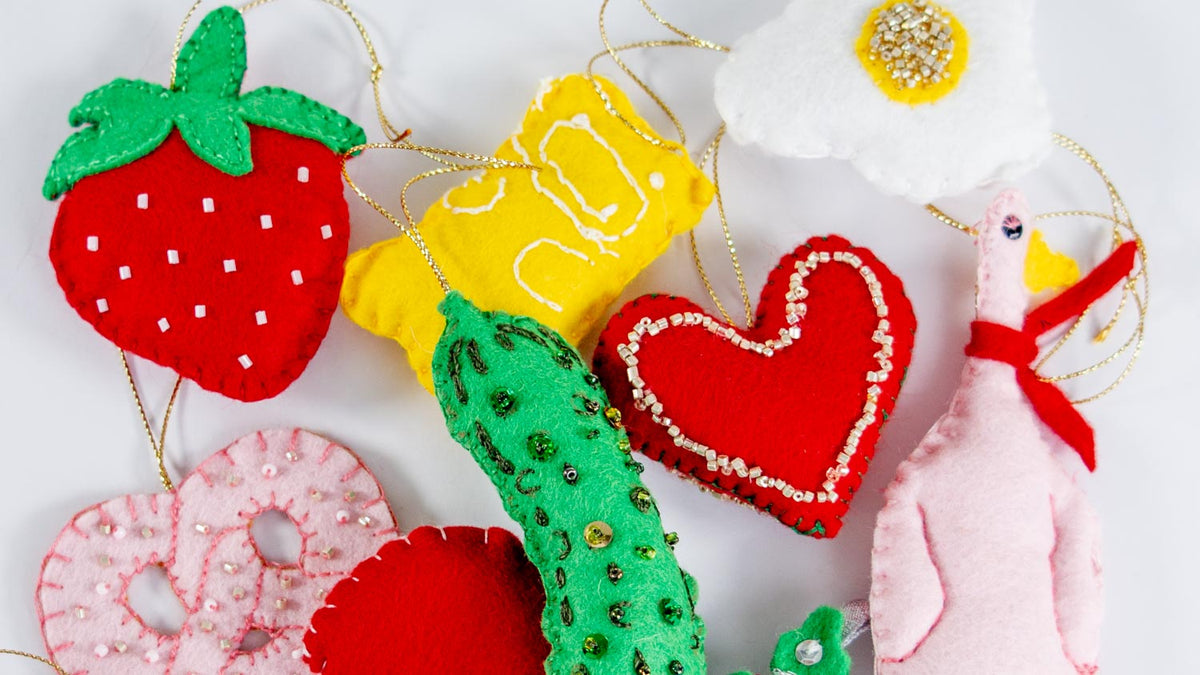 A top down image of felt Christmas decorations , featuring a strawberry, gummy bear, fried egg, pickle, heart, pretzel, mushroom and goose. 