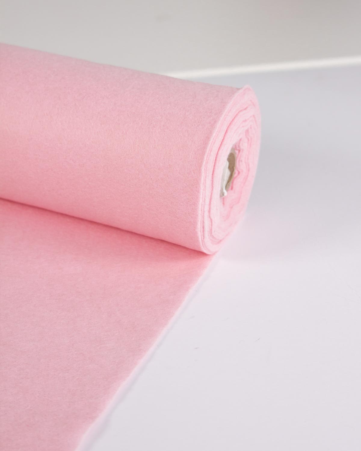 A roll of candy pink wool and viscose felt fabric, partially unrolled onto a plain white surface, showing off its bright colour. 
