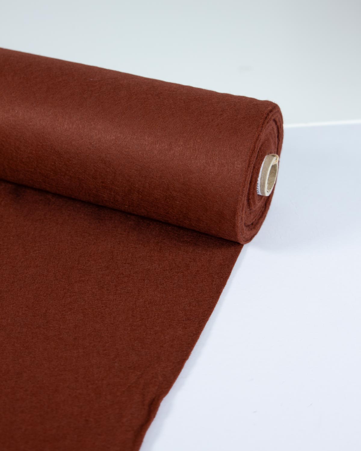 A roll of chestnut brown wool and viscose felt fabric, partially unrolled onto a plain white surface, showing its even colour and texture. 