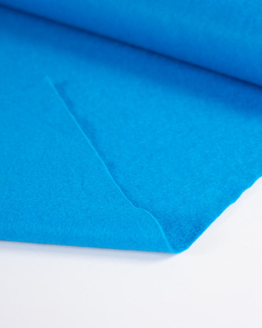 A piece of bright blue peacock wool and viscose felt fabric, folded to show its even matte surface on a white surface.