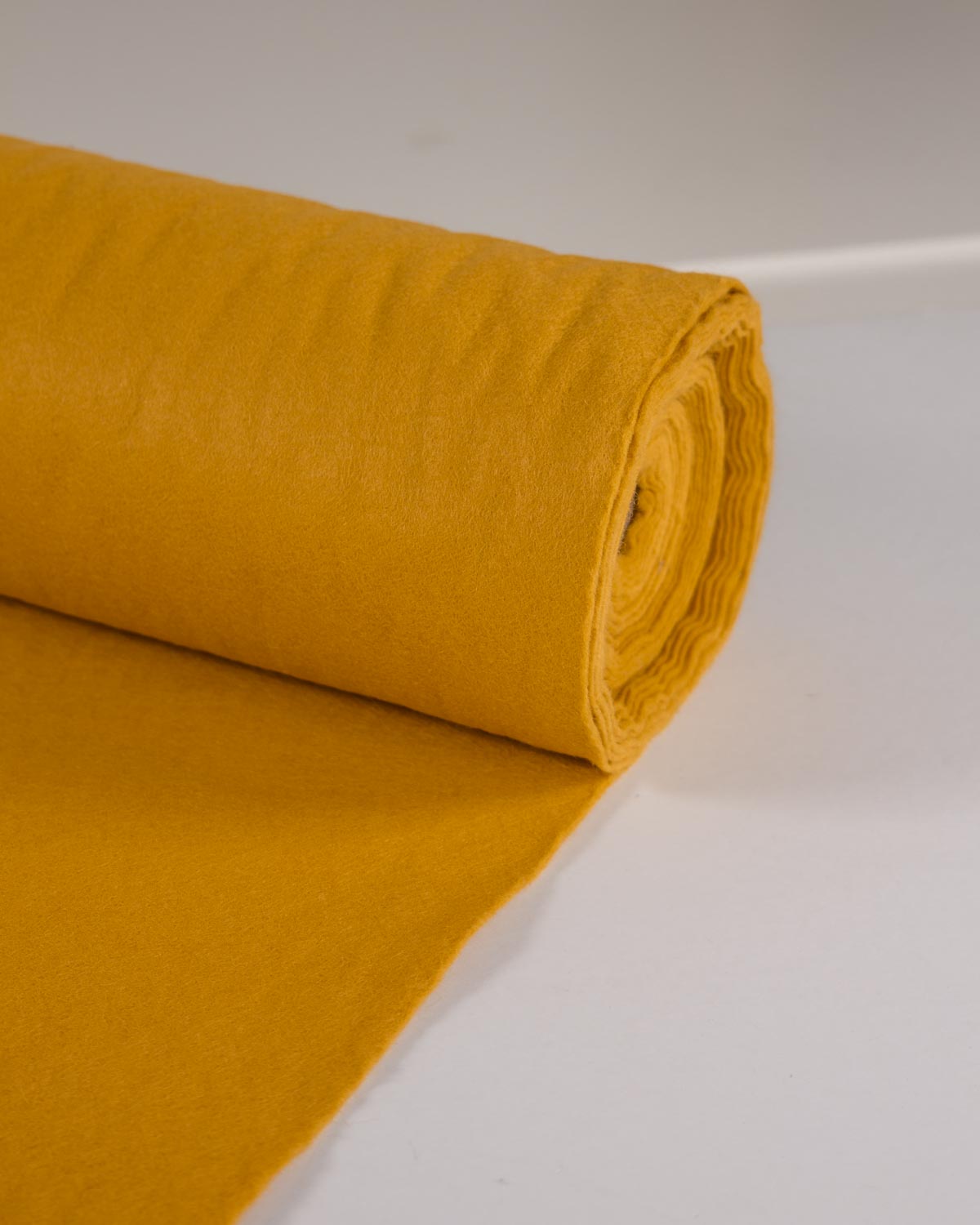 A roll of gold-yellow wool and viscose felt fabric unrolled onto a white surface. 