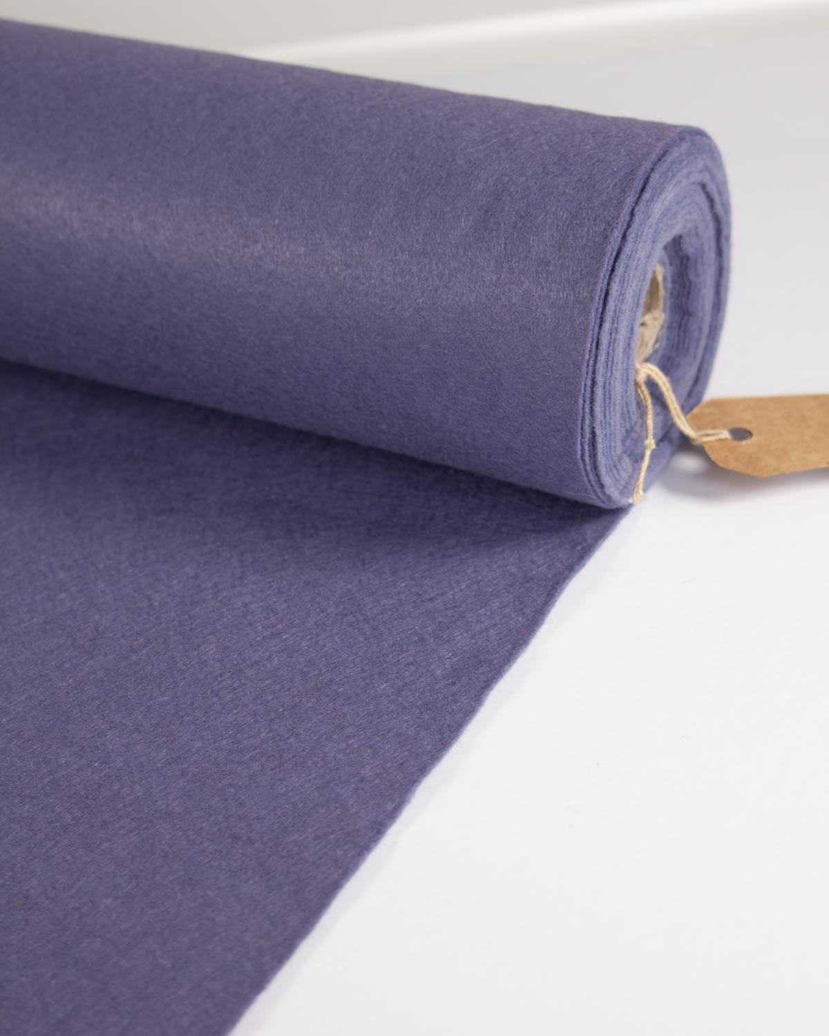 A roll of textured wool blend felt craft fabric in a muted periwinkle purple colour, unrolled slightly to show its weave, with a small kraft paper tag attached, set against a clean white background.