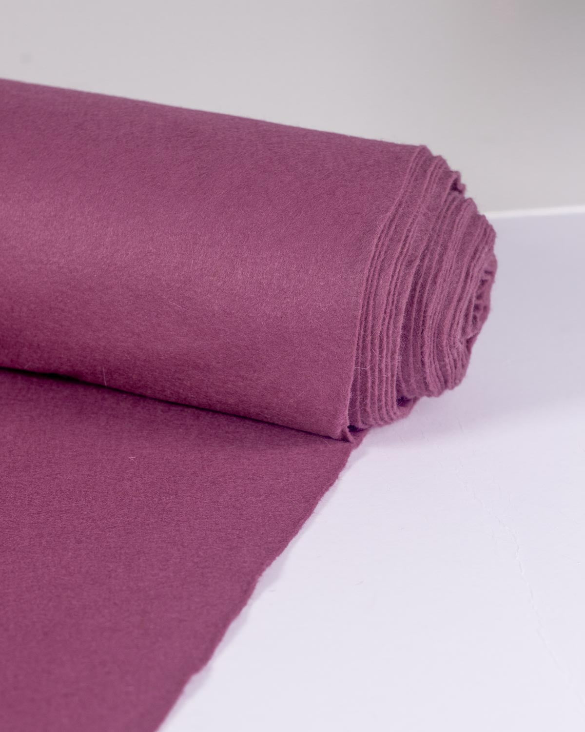 A roll of raspberry pink/purple wool and viscose felt fabric with a matching flat sheet unrolled, displayed on a clean white surface.