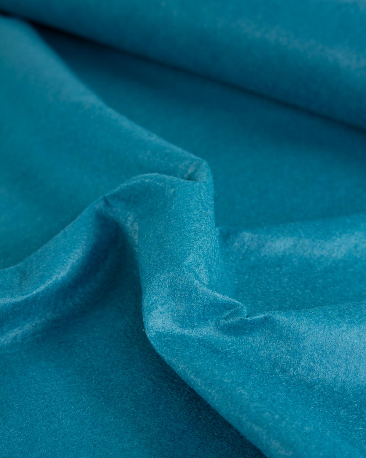 A close-up of draped teal blue wool and viscose felt fabric with a soft, velvety texture and gentle folds.