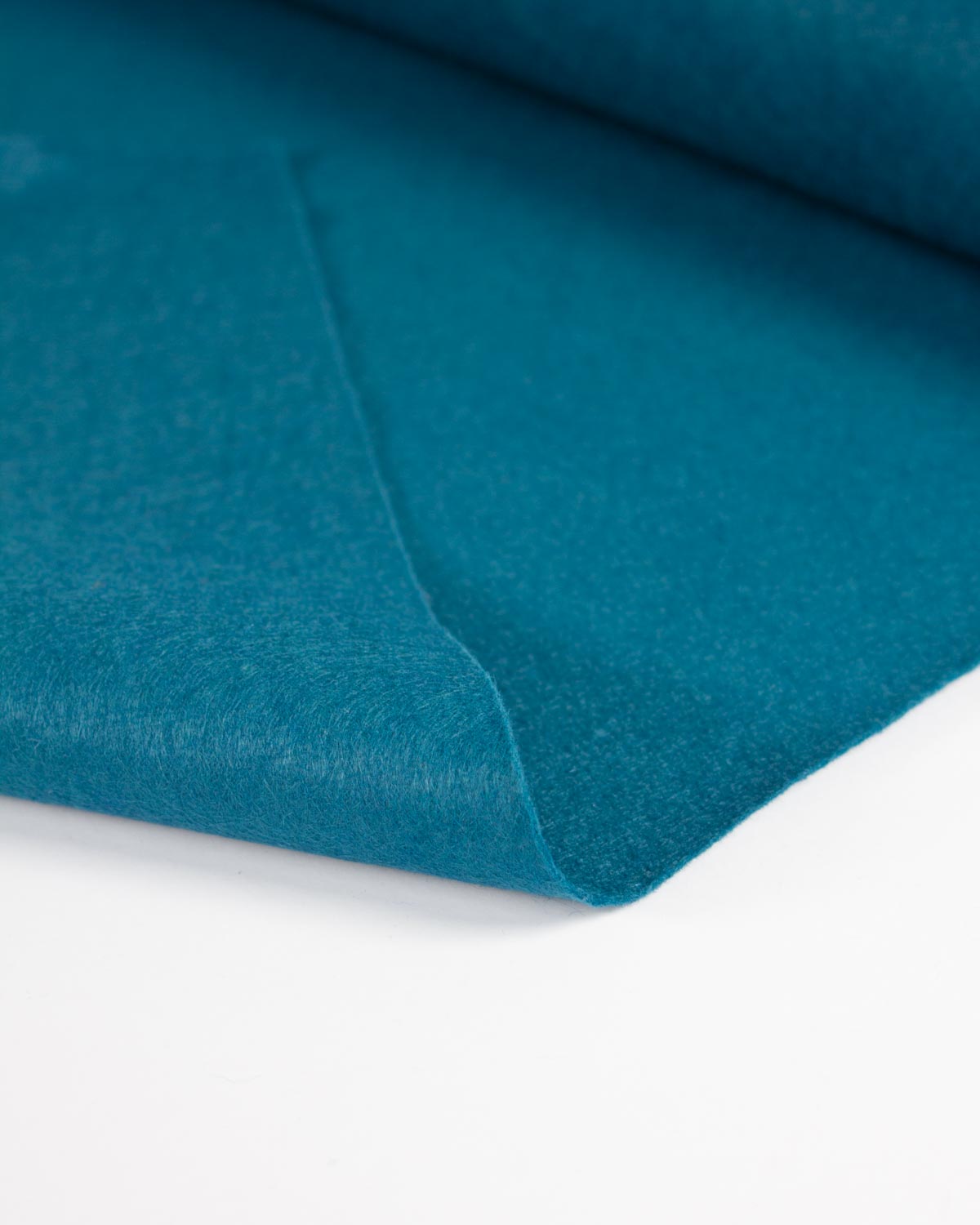 A close-up of a teal blue wool and viscose felt fabric, folded to show its bold matte texture on a flat white surface. 