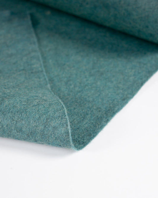 A close-up of a jade blue marl wool and viscose felt fabric folded on a plain white surface. 