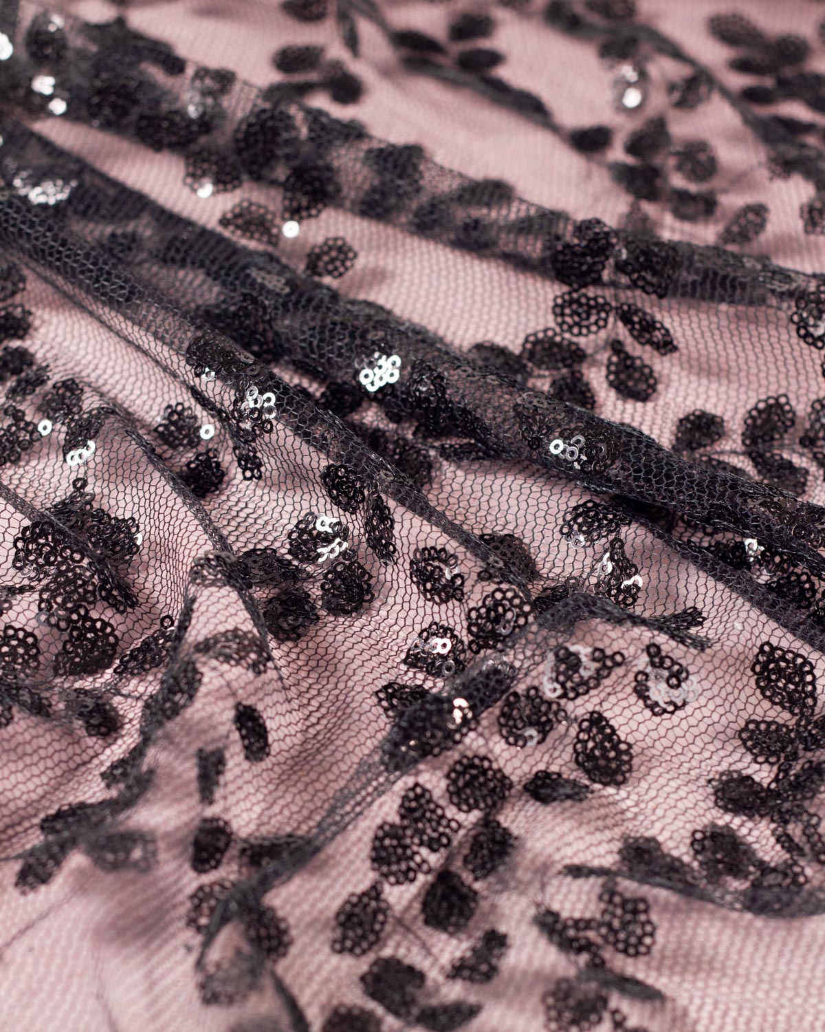 Close-up of a black sequined tulle fabric decorated with small black sequins in a floral leaf pattern on a dusty pink background. 