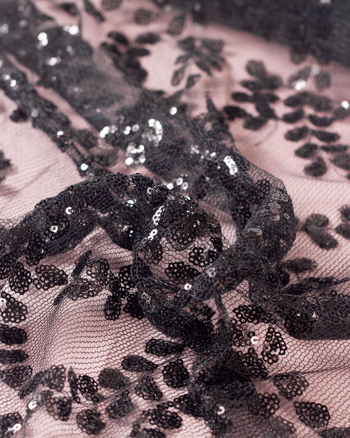 A gathered piece of black tulle fabric featuring a floral leaf design of small black sequins, set on a dusty pink backdrop. 
