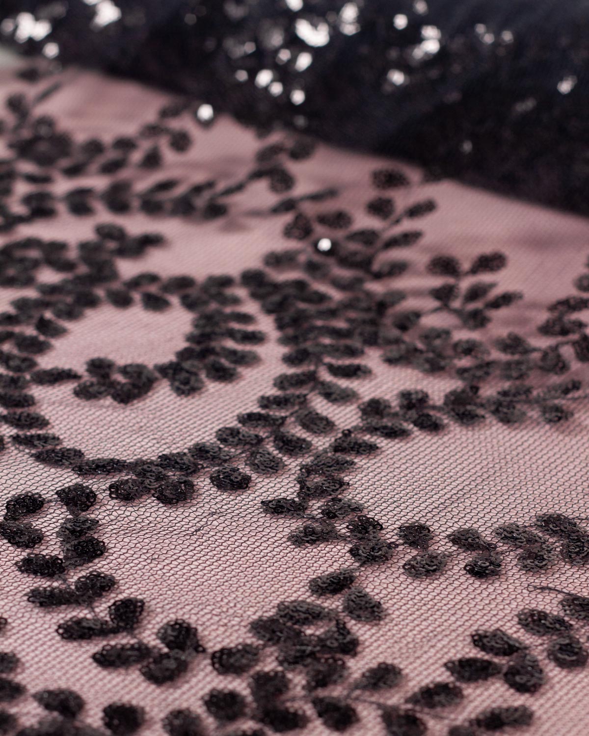 A piece of black tulle fabric with a floral leaf pattern of small black sequins unrolled onto a pink background. 