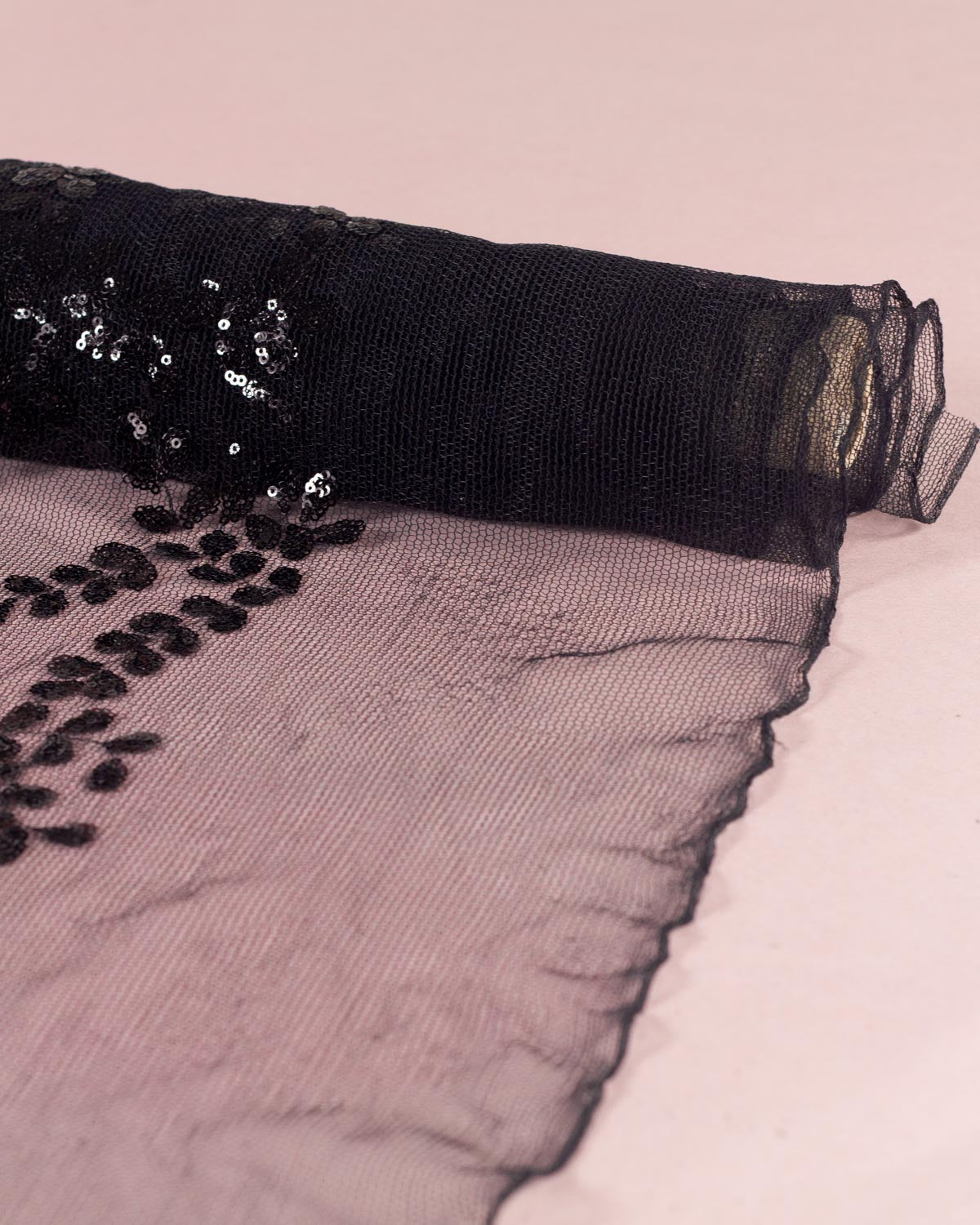 Black tulle fabric with sequined floral leaf applique partially unrolled on a light pink background.