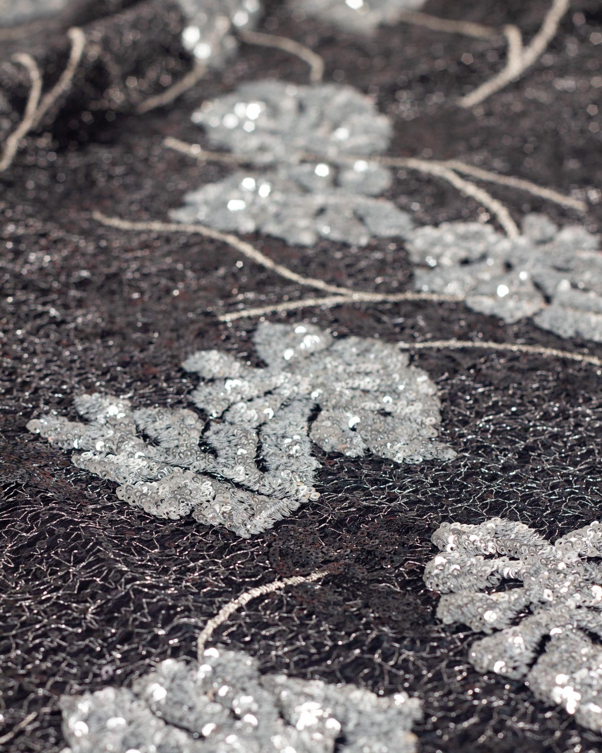 Close-up of a black cobweb tulle fabric with silver sequined monstera leaf motifs and thin metallic silver embroidered stems, with silver lurex running through the tulle. 