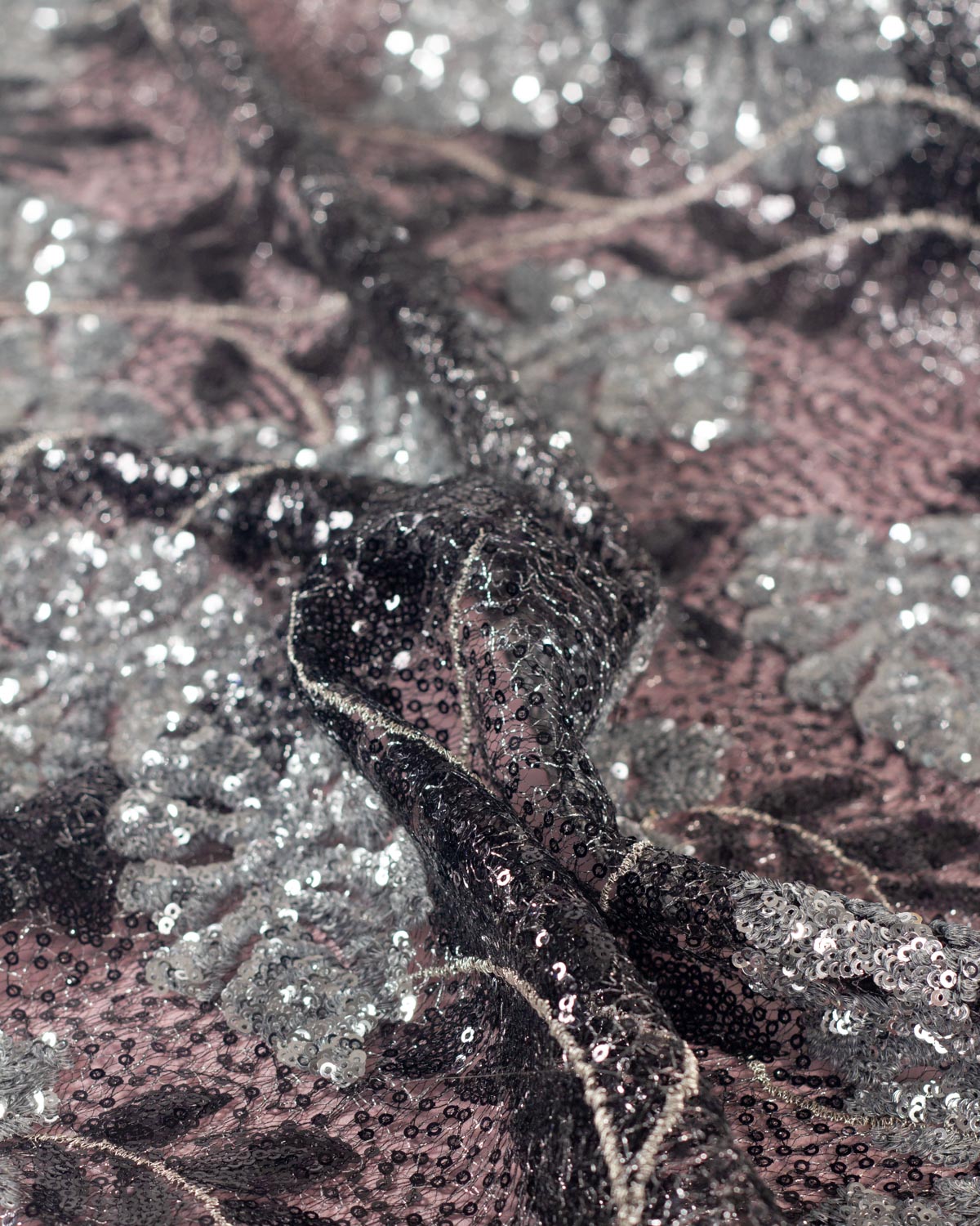 Close-up of a sequined cobweb black tulle fabric with a silver sequined monstera leaf pattern, featuring black sequins and silver lurex woven through over a dusty rose backdrop.