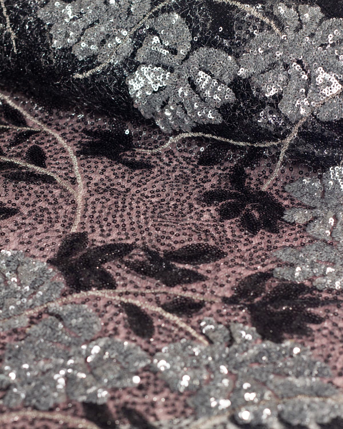 A partially unrolled of black cobweb tulle fabric with silver lurex woven in featuring a design of silver sequined monstera leaves, black sequined leaves and silver embroidered stems, set on a dusty pink backdrop. 