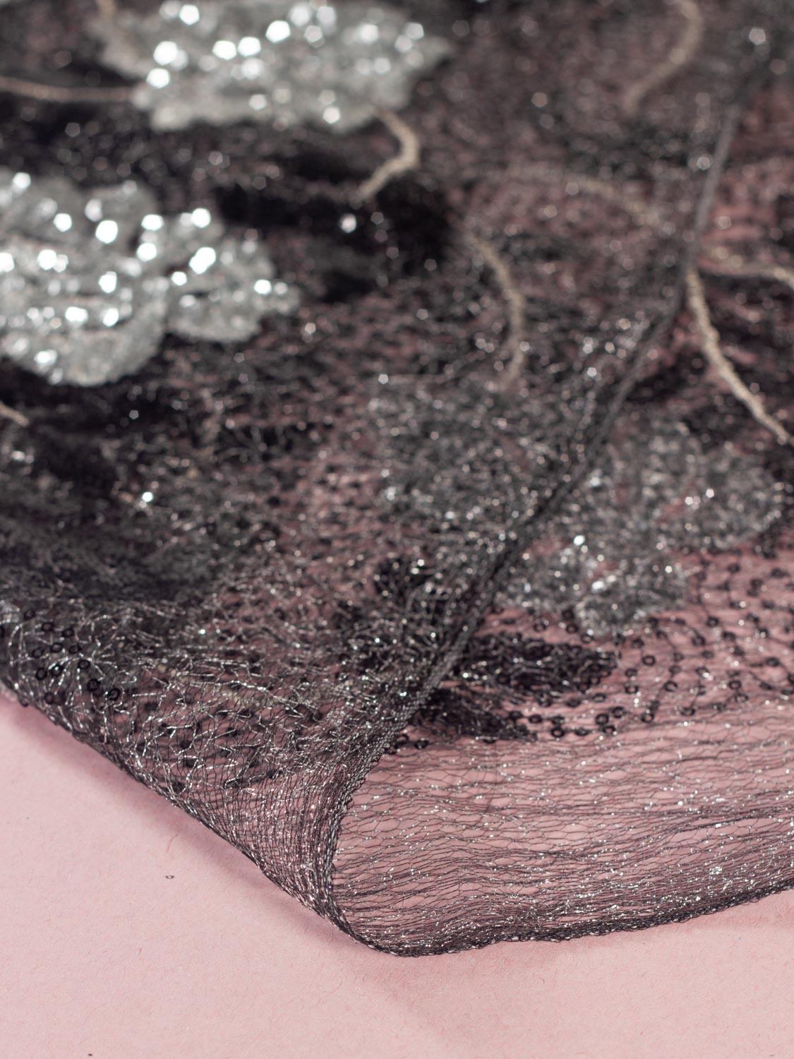 Close-up of a sheer black tulle fabric with silver sequins and silver lurex woven through , draped over a light pink surface.
