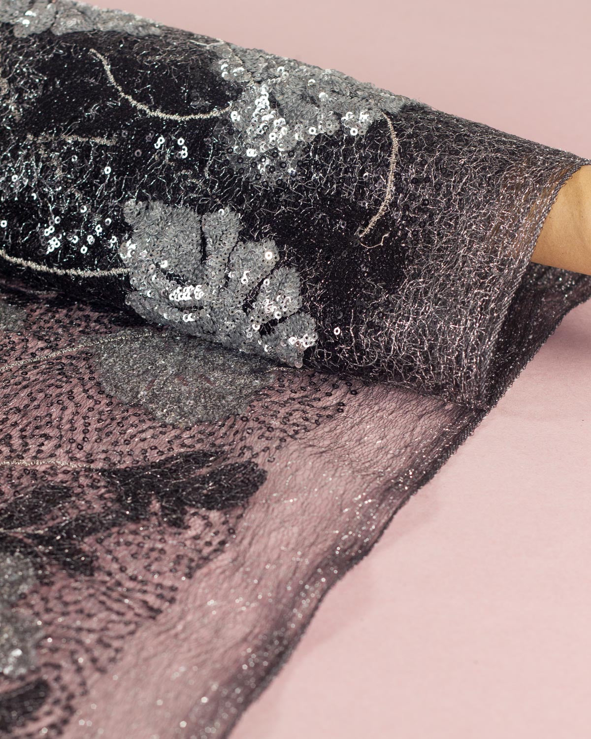 A roll of of black cobweb tulle fabric with silver lurex woven through, partially unrolled onto a pink surface, showing its texture and densely silver sequined pattern of monstera leaves, black leaves and silver embroidery. 