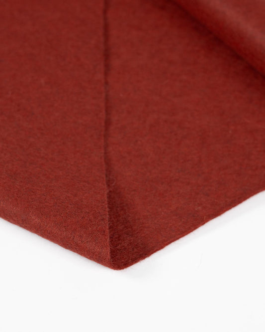 A close up of a wool and viscose felt fabric in a terracotta red/orange marl, folded to show its texture and marled finish on a white surface. 