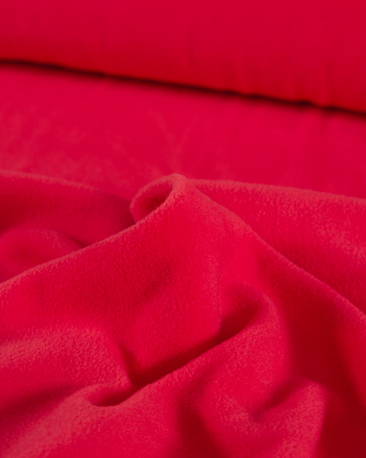 A close-up of bright red plush fleece fabric with soft folds and a smooth, slightly lighter red band across the top edge.