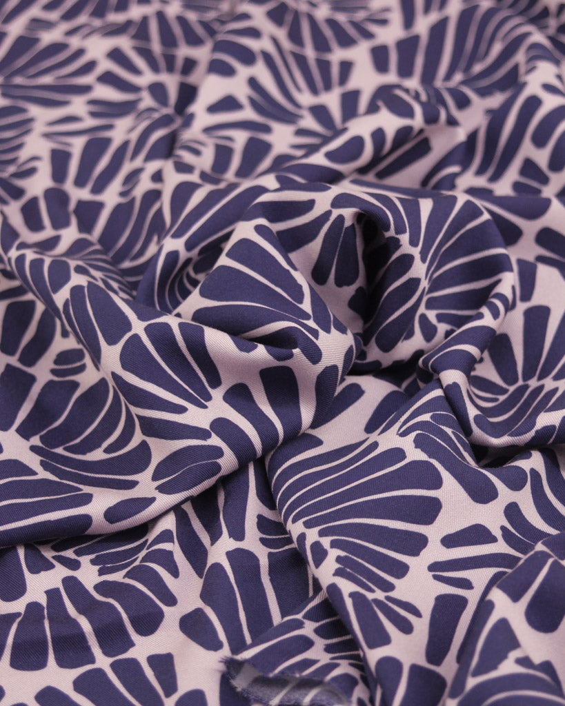 Close up of viscose twill fabric featuring an all-over palm fan motif in deep, rich purple on a soft dusty mauve ground