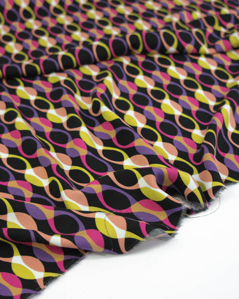 Colourful viscose twill fabric with a pattern of interlocking shapes of pink, yellow and purple on a black background