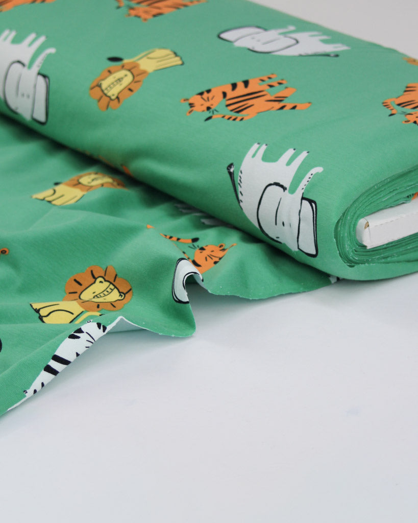 Roll of green cotton jersey fabric with cartoon animal prints on a white background. Featuring lions, tigers and elephants.