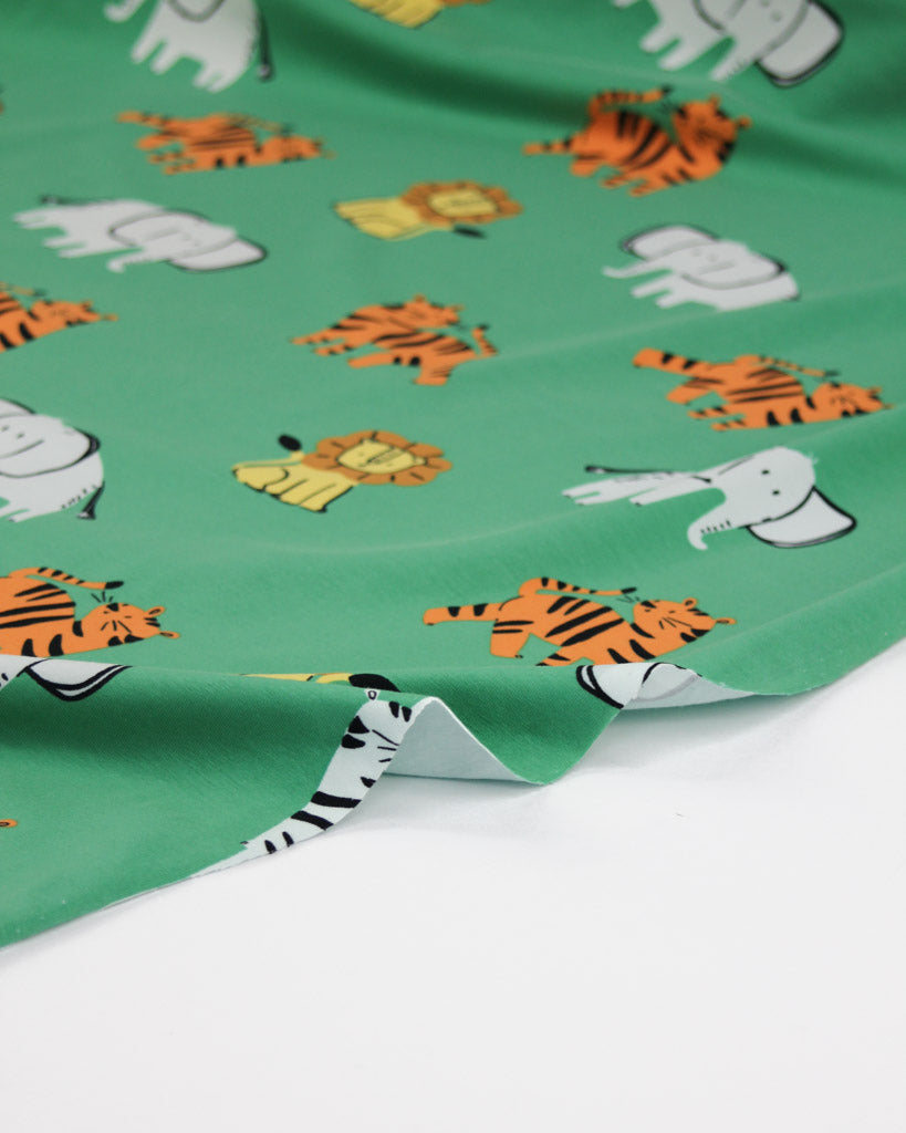 Roll of green cotton jersey fabric with cartoon animal prints on a white background, featuring lions, tigers and elephants.
