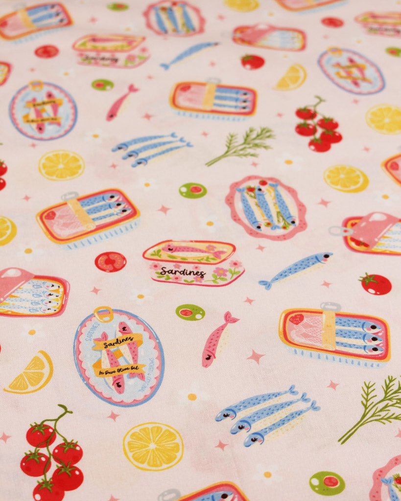 Cotton fabric with a colourful pattern of sardine tins, fish, and vegetables on a light pink background.