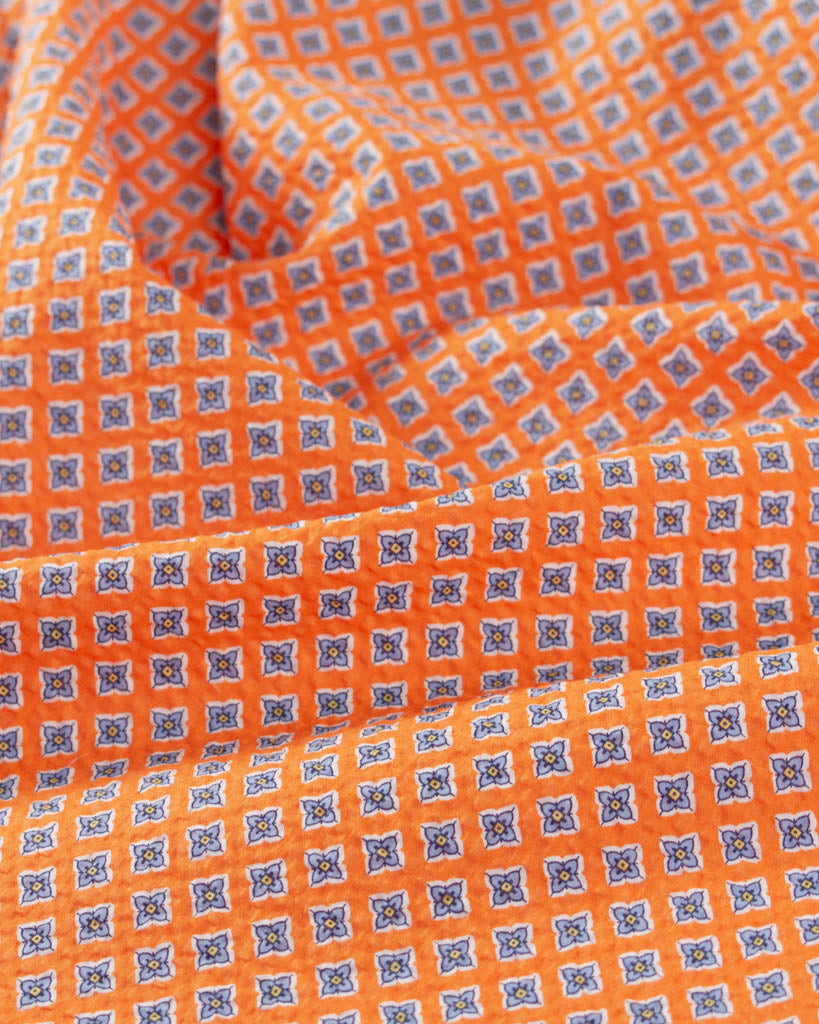 Rippled view of mango orange cotton seersucker fabric with small four-petal floral tile print