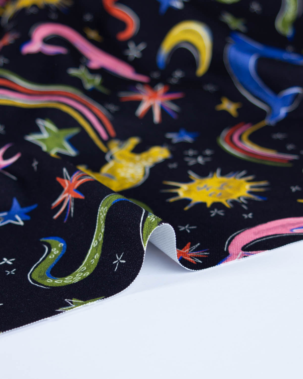 A close-up of a black denim fabric with a colourful cosmic print featuring stars, rainbows, and planets, with the underside showing a white backing.