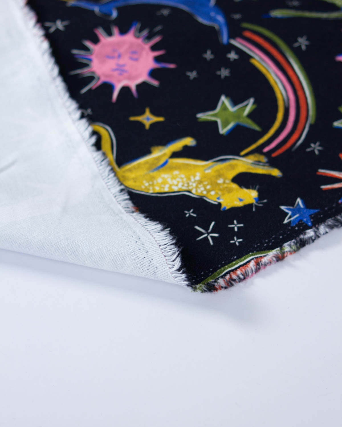 A close-up of a denim fabric, showing a dark navy blue space-themed print with a yellow cat, rainbow, stars, and sun.
