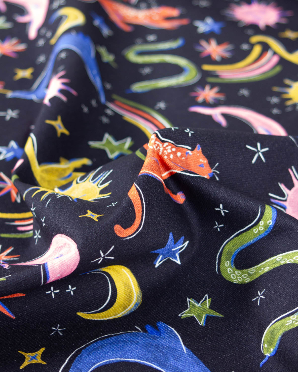 A close-up of a black denim fabric with a colourful cosmic print featuring stars, crescent moons, rainbows, and whimsical space-themed illustrations.