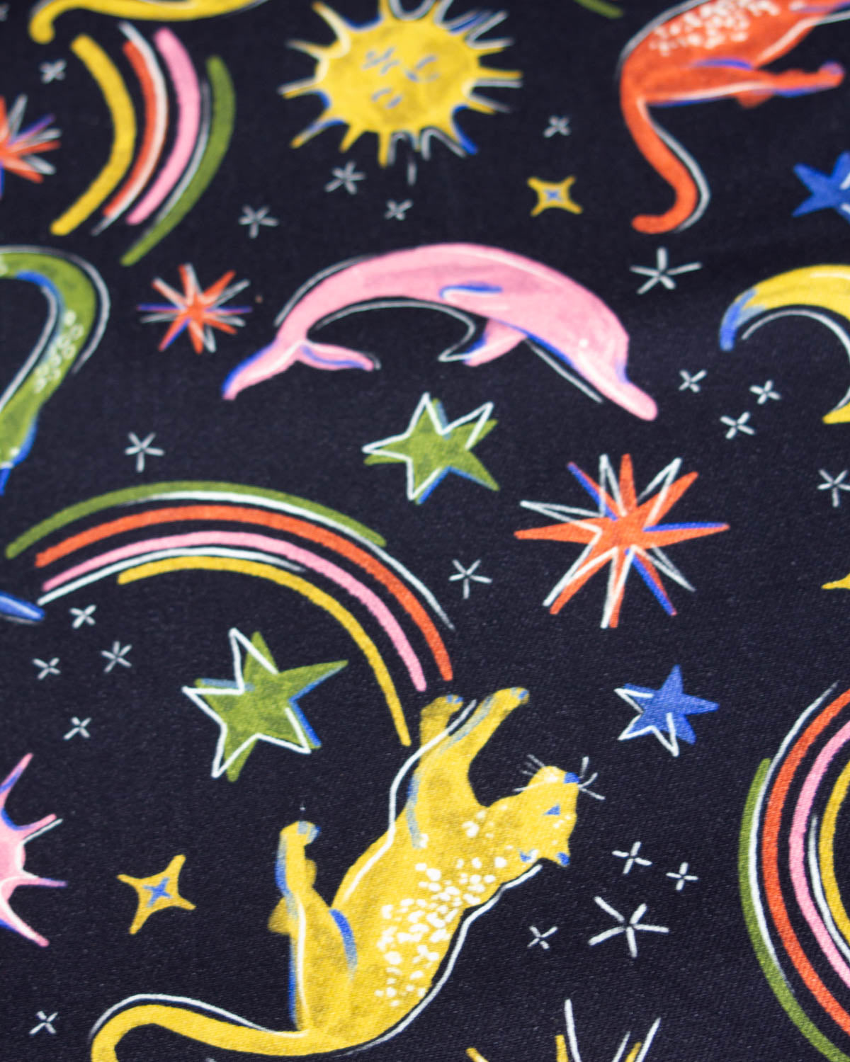A close-up of a denim fabric featuring a black background decorated with colorful illustrations of a yellow cat, pink dolphin, yellow sun, green stars, and rainbow arcs.