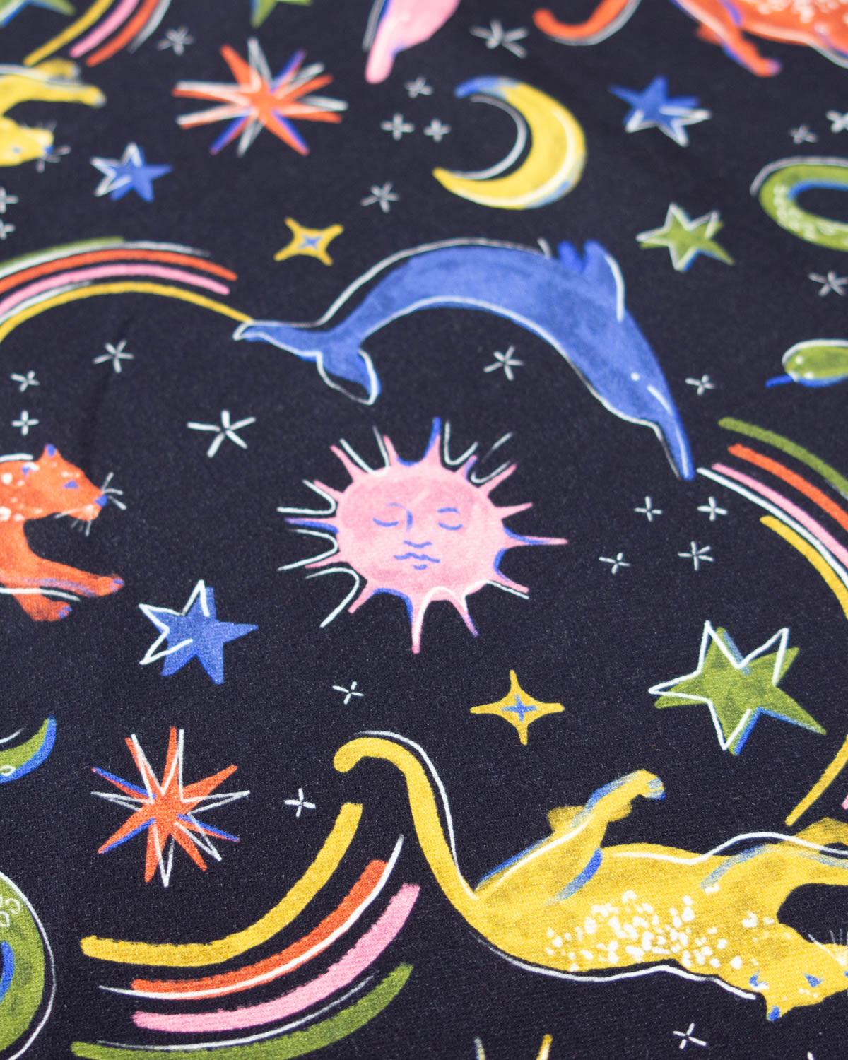 A close-up of a denim fabric featuring a whimsical space theme with a pink sun, blue dolphin, yellow cat, colorful stars, and rainbows on a dark navy background.