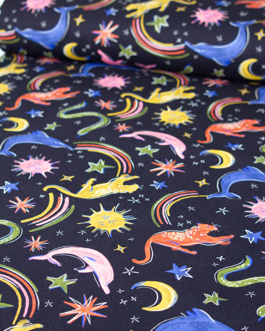 A close-up of a black denim fabric with a colourful whimsical print featuring stars, rainbows, dolphins, crocodiles, cats, and moons in bright pink, yellow, orange, green, and blue.