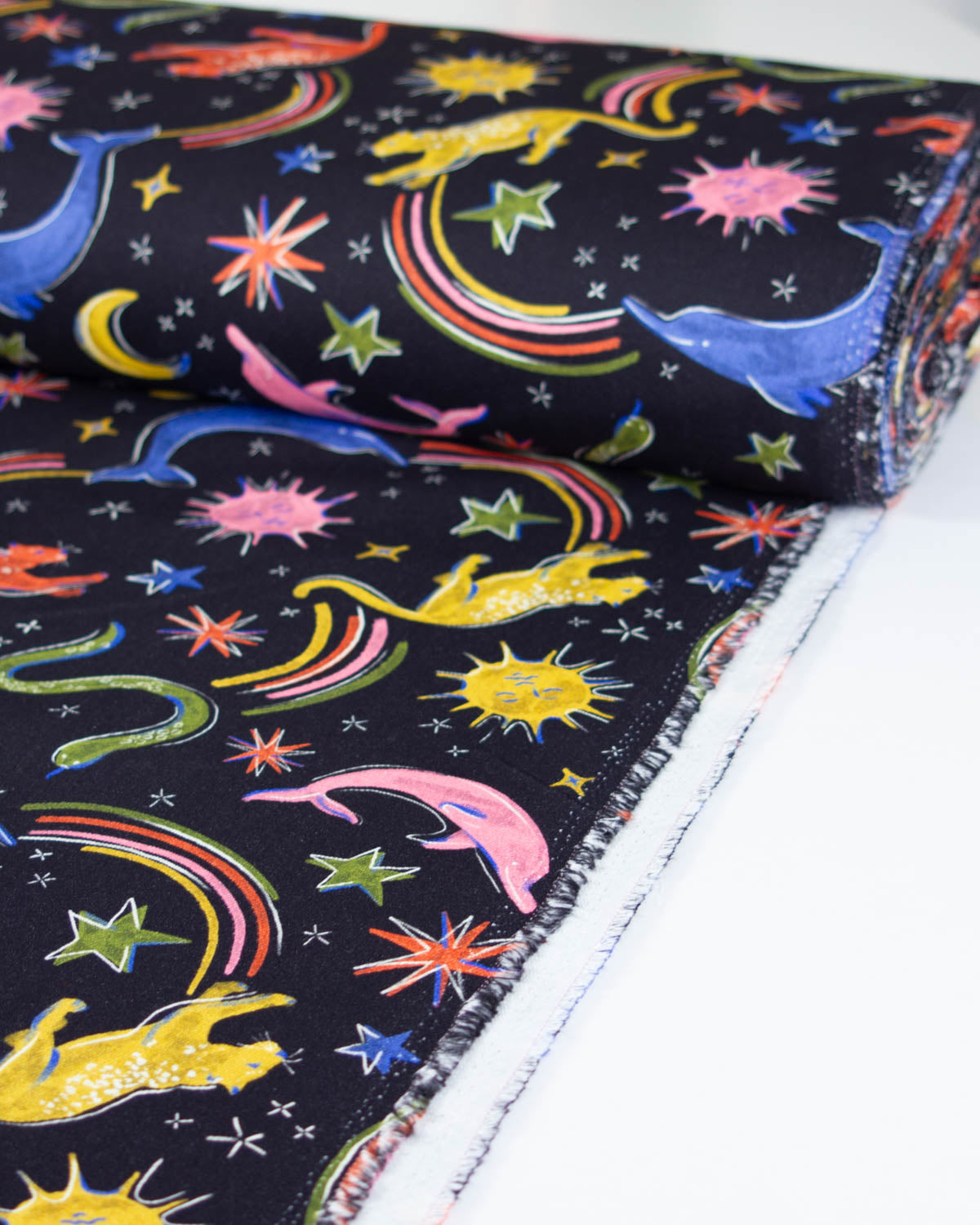 A close-up of a rolled denim fabric with a black background and a colourful print of cartoon animals, stars, and rainbows, shown on a white surface.
