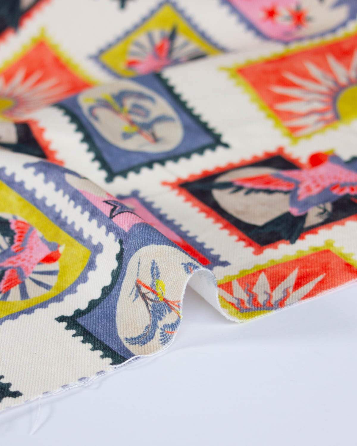 A close-up of a printed cotton denim fabric with a colorful postage stamp pattern, featuring palm trees, blue birds, and yellow sunburst motifs on a white background.